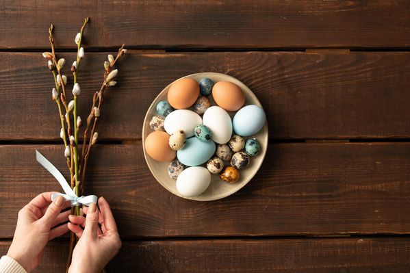 Top view of a beige plate filled with Easter quail and chicken eggs and a bouquet of willow twigs tied with a blue ribbon that the hands touch