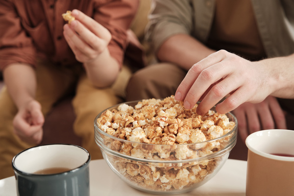 Popcorn in a glass bowl standing on a table with mugs of tea is taken by people sitting on the sofa