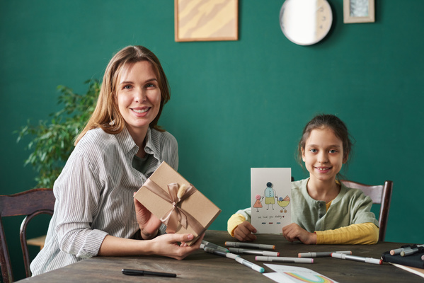 A woman in a striped blouse with a gift in a craft box decorated with a ribbon and her daughter with tidied up hair with a homemade postcard with an illustration sitting at a table on which markers are scattered