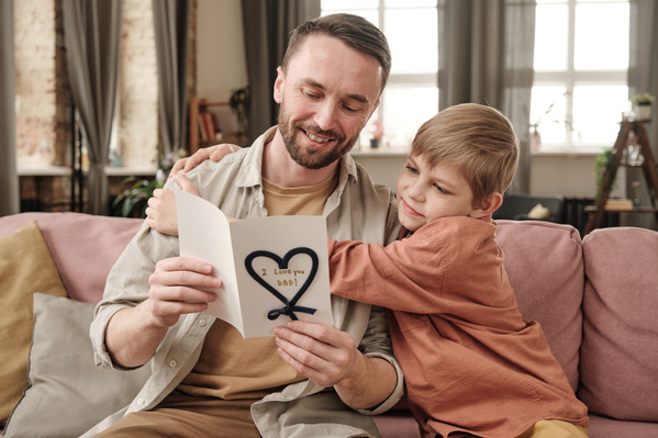 A boy in a brick-colored shirt hugging his dad reading congratulations in a homemade Fathers Day card while sitting on the couch