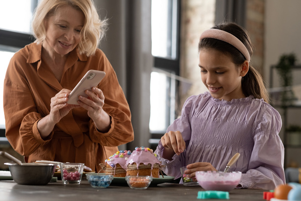 A grandma with blonde hair takes a photo on her phone of her granddaughter on her smartphone in a pink blouse decorating glazed Easter cakes with colorful sweets