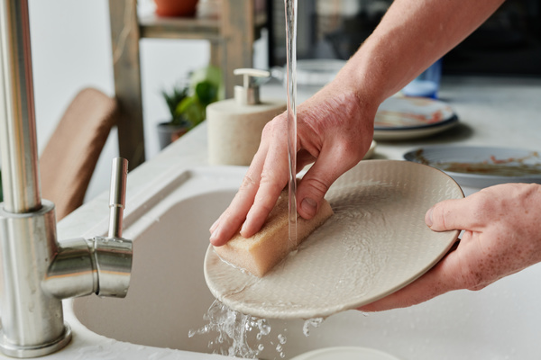 A beige dish is cleaned under a stream of water with a light-colored sponge in a white sink with dirty dishes