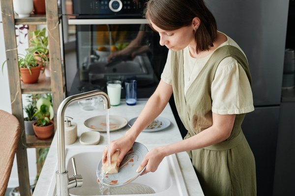 A woman with dark hair washing a gray plate under a stream of water with a light-colored sponge in a white sink with dirty dishes