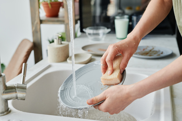A gray plate is washed under a stream of water with a light-colored sponge in a white sink with dirty dishes