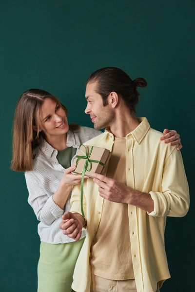 A wife in a striped blouse presenting a gift in a craft box decorated with a green ribbon to her husband in a yellow shirt standing against an emerald background