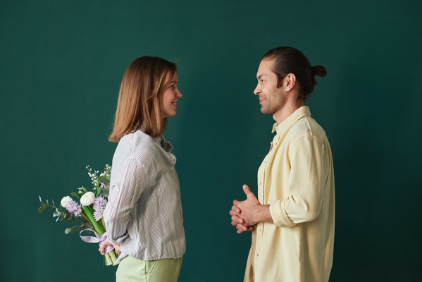 A wife in green trousers hiding a bouquet of flowers behind her back congratulating her husband in a yellow shirt standing against an emerald-hued background