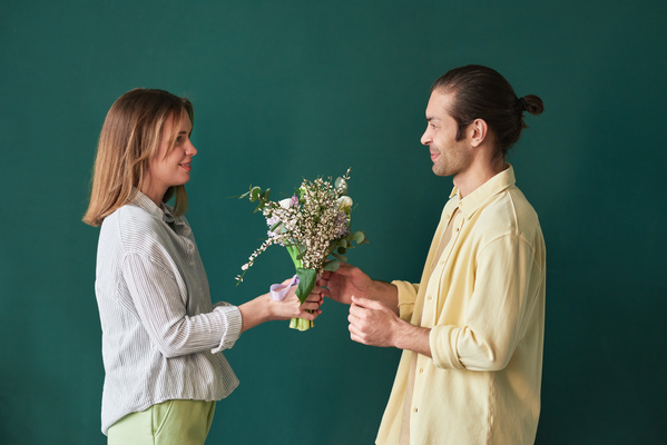 A woman in green trousers giving a bouquet of flowers to her husband in a yellow shirt standing against an emerald background