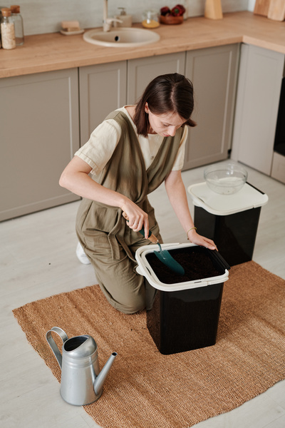 A woman with short dark hair loosening the fertilized soil in a black bucket with a green garden shovel sitting on the kitchen floor