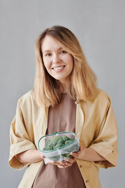 A woman with blonde hair dressed in light-colored clothes smiling holds fresh broccoli in a reusable vegetable bag
