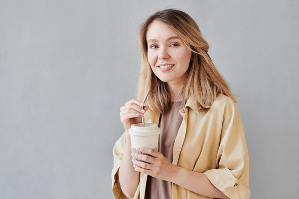 A woman with blonde hair dressed in beige clothes holds an eco-friendly white cup with an iron straw