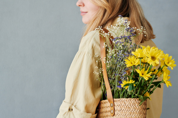 A blonde woman dressed in light clothes with a straw shopper of flowers hanging on her shoulder