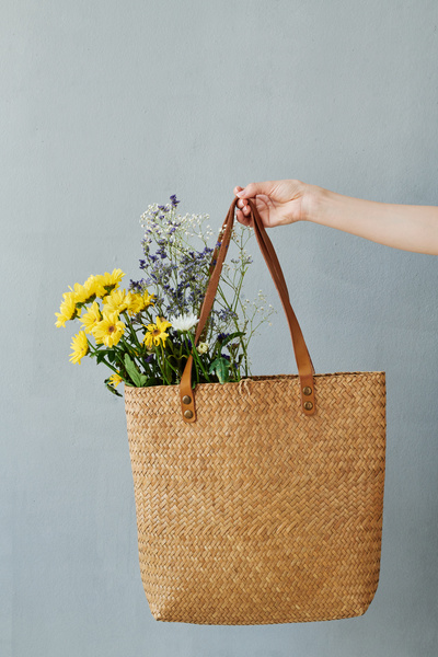 A wicker straw shopper with bright flowers in a womans hand on a light background