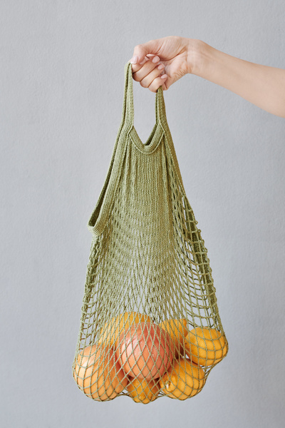 A green organic string bag filled with citrus fruits is held in the hand on a light background