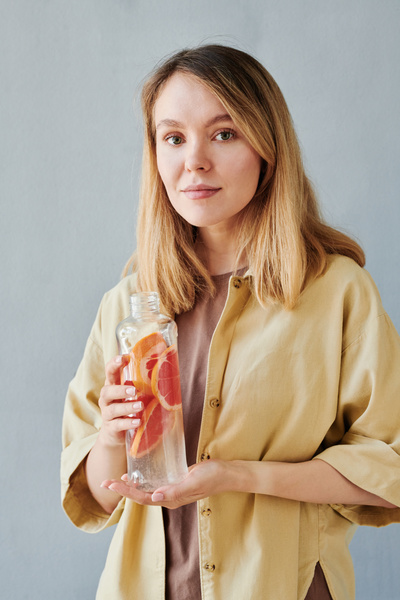 A blonde woman dressed in a beige shirt with a reusable bottle of drink with grapefruit slices
