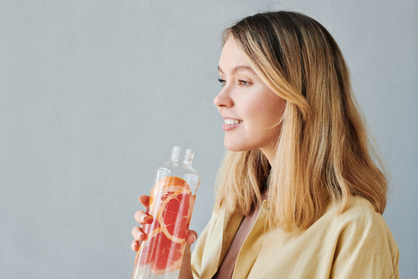A blonde woman in a beige shirt smiling looks away holding a transparent bottle with a grapefruit drink