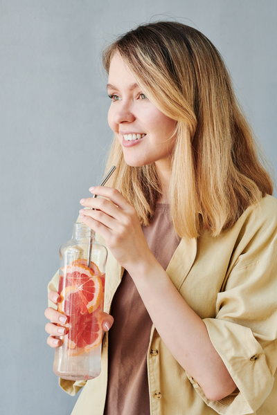 A woman with blonde hair in a beige shirt holds a transparent bottle with grapefruit water and a metal eco straw