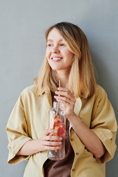 A woman with blonde hair in a beige shirt with a transparent bottle of grapefruit water and a metal eco straw