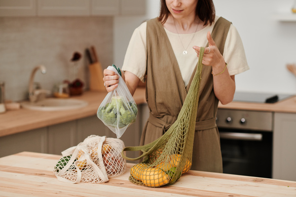 A woman with dark short hair holding organic bags with lemons and broccoli standing at the kitchen table with a white cotton shopping bag with groceries on it
