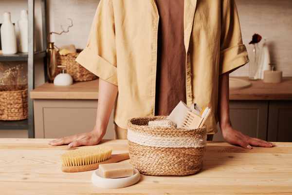 A woman dressed in beige-colored clothes standing behind a wooden table with a wicker eco-friendly basket of toiletries on it