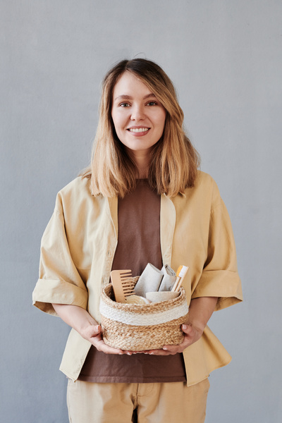 A woman with blonde hair dressed in beige clothes smiling holding a wicker basket with toiletries