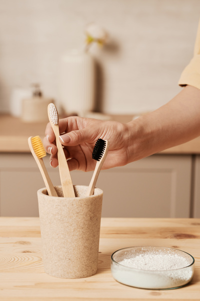 A woman taking a wooden eco-friendly toothbrush with white bristles from a beige toothbrush holder on a table with a glass petri dish with tooth powder