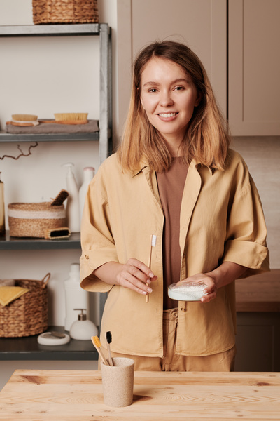 A woman with blonde hair dressed in beige clothes smiling holds an eco-friendly toothbrush and tooth powder in a glass petri dish