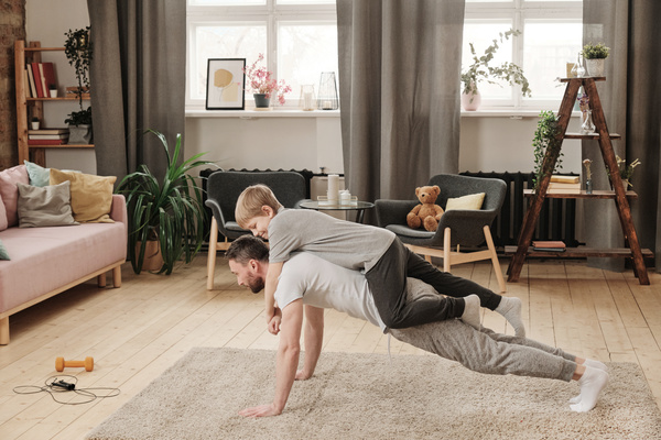 A blond-haired boy in a gray T-shirt is on the back of his dad doing push-ups on a light carpet weighing him down