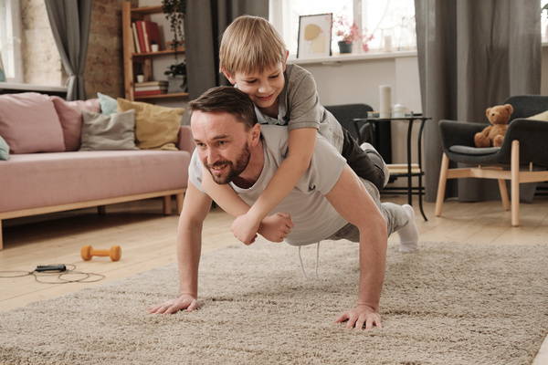 A man in a sportswear doing push-ups with a blond son on his back in a gray T-shirt on the carpet in the living room