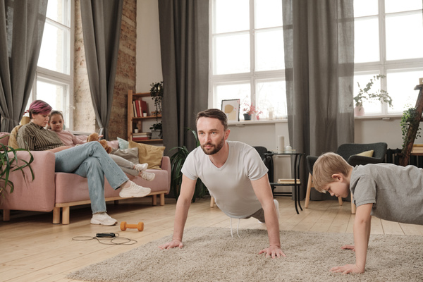 A man with a beard dressed in a white T-shirt doing push-ups on the carpet in the living room with a blond son in a gray T-shirt