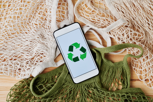 A phone with the image of a green garbage recycling sign on the screen on a white organic string bag