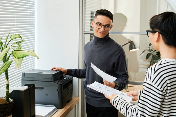 An office worker with dark hair dressed in a gray sweater makes a photocopy looking at a colleague in a striped longsleeve