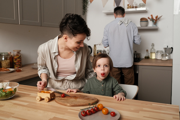 A little boy playing with a tomato while cooking a salad with his dark-haired mom