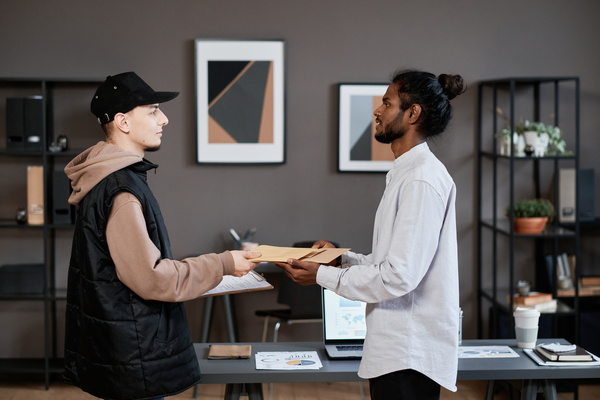 A courier in a black cap giving documents in a craft package to a businessman with his hair in a bun and dressed in a white shirt in the office