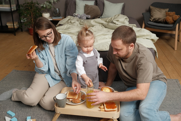 A family consisting of a mom with short hair her husband with a beard and their daughter in a gray jumpsuit having breakfast with toastes with jam and orange juice in the bedroom