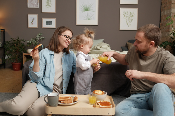 A man in a beige T-shirt pouring orange juice to his little blonde daughter and mom eating a toast with jam at home breakfast