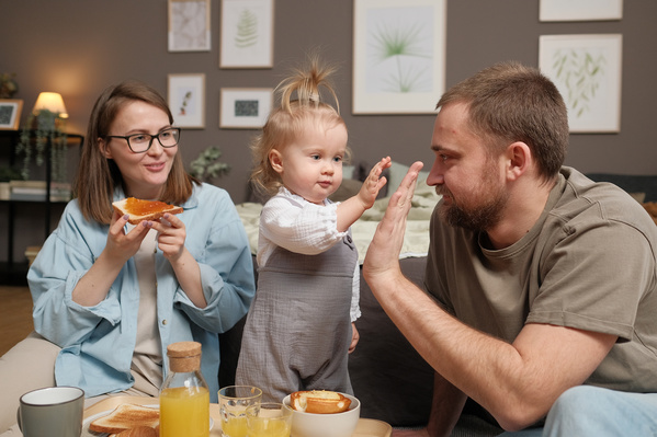 A blonde toddler girl high-fiving her dad and mom eating toast with jam at a family breakfast