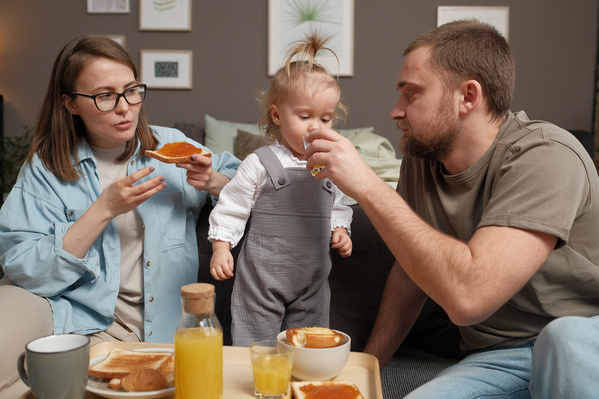 A man in a beige T-shirt treating his little blonde daughter with orange juice and a mother eating toast with jam at a family breakfast