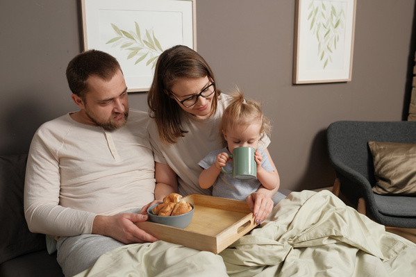 Parents in light pajamas sitting in bed with breakfast on a wooden tray and their daughter drinking tea from a green mug