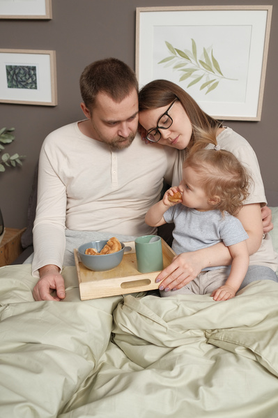 Parents in light pajamas tenderly looking at their daughter having breakfast with croissants in bed