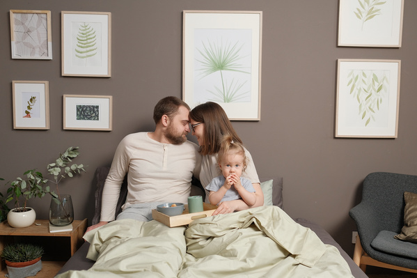 Hugging parents in light pajamas and their daughter having breakfast with croissants in bed