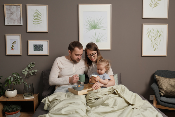 Parents in light pajamas and their daughter sitting in bed with breakfast on a tray and mugs
