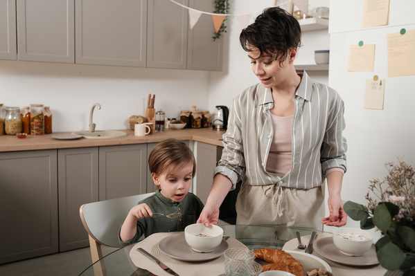 A woman with short dark hair serving porridge in a white bowl to her little son sitting at the breakfast table