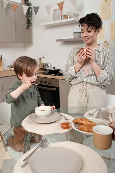A dark-haired woman with a mug in her hands looking at her little son having breakfast porridge at a glass table with croissants on it