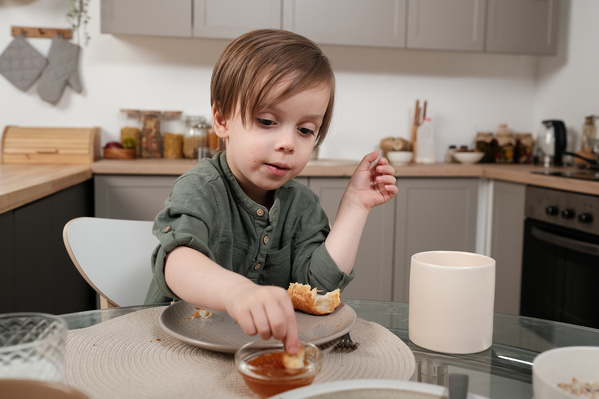 A little blond boy dipping a piece of croissant into jam at breakfast