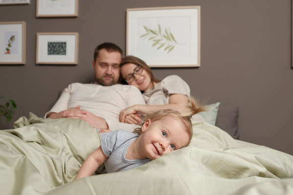 A blonde toddler girl in gray T-shirt lying in bed with her relaxed parents