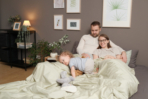 Parents in light pajamas and their little daughter with soft toys in bed