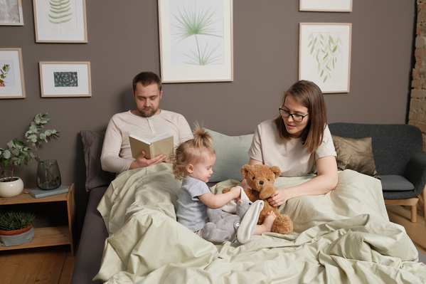 Mom in glasses playing with her little blonde daughter with a stuffed toy and dad in pajamas reading a book in bed