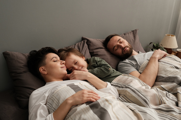 A boy in green pajamas sleeping with his dark-haired mother and father in a gray T-shirt in bed