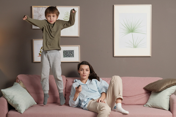 A woman in a light shirt watching TV and her son in pajamas jumping on a pink sofa