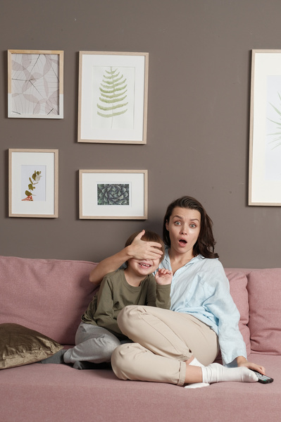 A woman in light clothes with a surprised expression covering her little sons eyes with her hand while watching a movie on a pink sofa